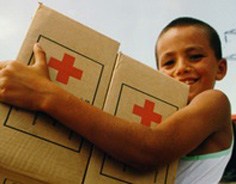 A boy carries two cardboard boxes with red crosses on the side, smiling.