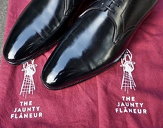 A close-up shot of the toes of a pair of black dress shoes on a cloth that has "The Jaunty Flaneur" printed onto it twice.
