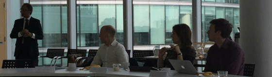 Three people sit and one man stands at a boardroom desk.