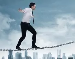 A man in a shirt, tie and suit pants walks a tightrope made of metal chain links, with a city skyline in the background.