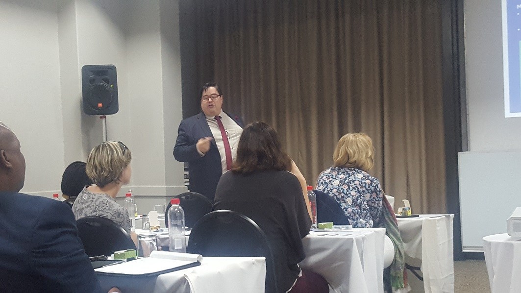 A man in a suit and tie addresses a group of people sitting at desks.