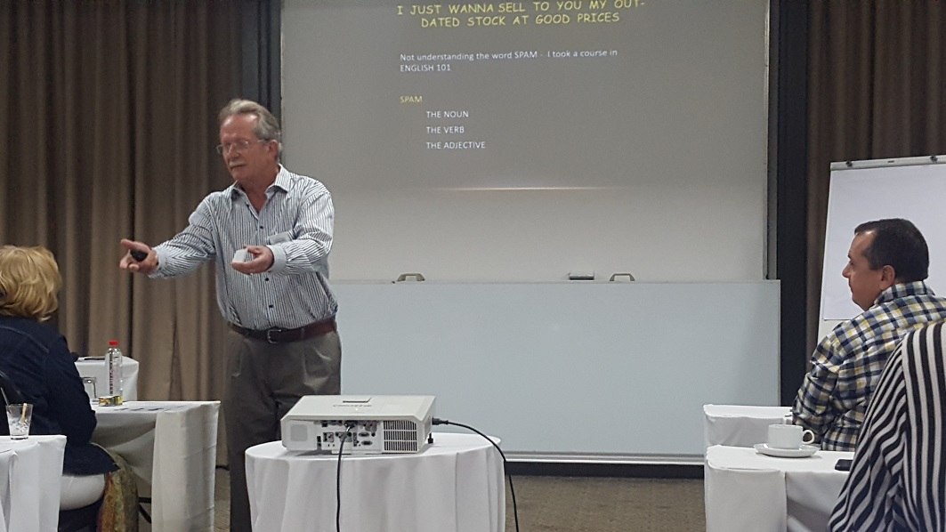 A man in a shirt addresses a group of people sitting at desks.