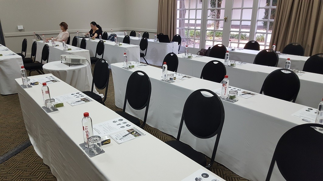 A room set up for a lecture with four rows of black chairs and tables covered in white cloths; the chairs are mostly empty.