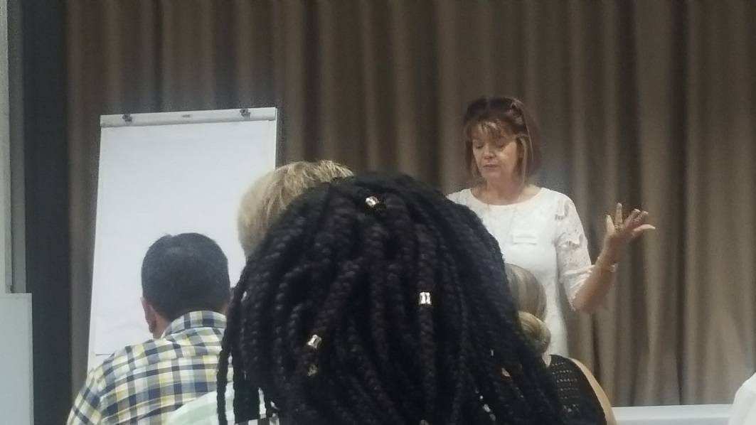 A woman in a white shirt addresses three people, seated in rows.