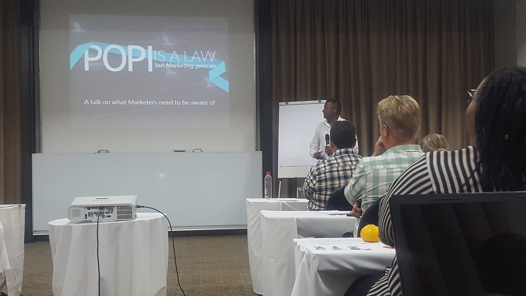 A group of people sit at tables, facing a screen that says "POPI"