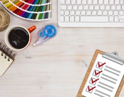 A birds-eye-view of a desk showing a keyboard, cup of coffee, colour swatches, tape dispenser, and checklist on a clipboard.