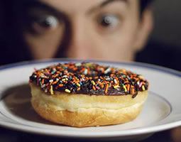 A close up of a donut with chocolate icing and rainbow sprinkles on a plate, with a man's wide-eyed face in the background.