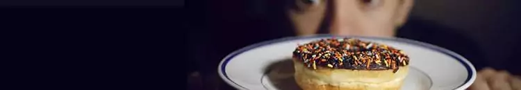 A close up of a donut with chocolate icing and rainbow sprinkles on a plate, with a man's wide-eyed face in the background.