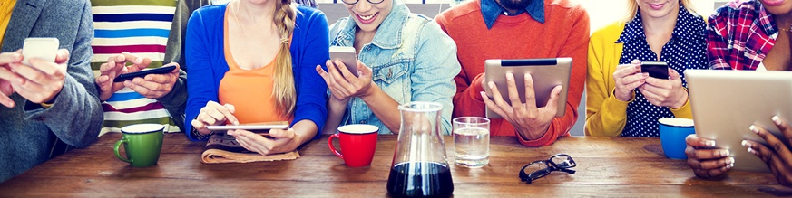 7 people sit in a row at a table, each wearing colourful clothing and holding a tablet or smartphone.