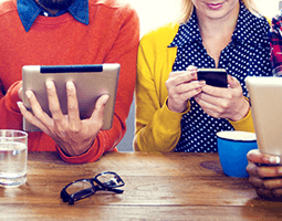 7 people sit in a row at a table, each wearing colourful clothing and holding a tablet or smartphone.