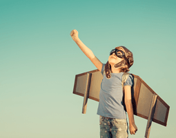 A child wearing a flying hat and goggles and strapped on plane wings makes a fist and looks up at the sky.