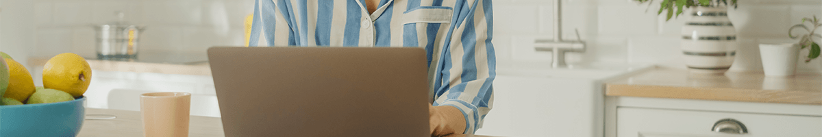 A woman sits behind a laptop in a striped blue shirt with a kitchen in the background.