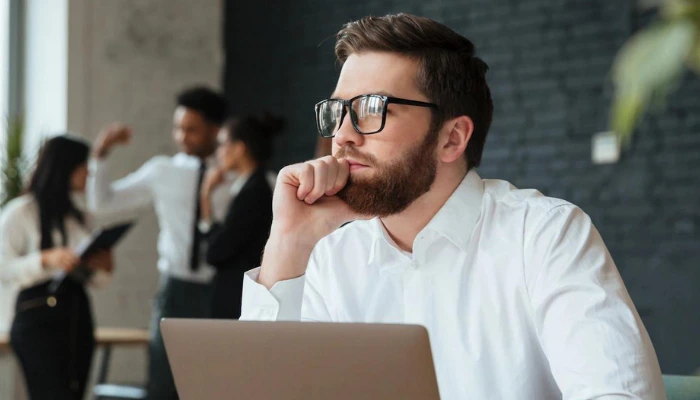 A man in a white shirt and glasses looks into the distance over an open laptop; three people in business attire talk in the background.