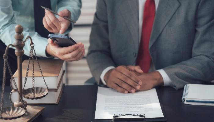 Two business professionals in suits reviewing information on a smartphone at a desk with legal documents, a notepad, and scales of justice.