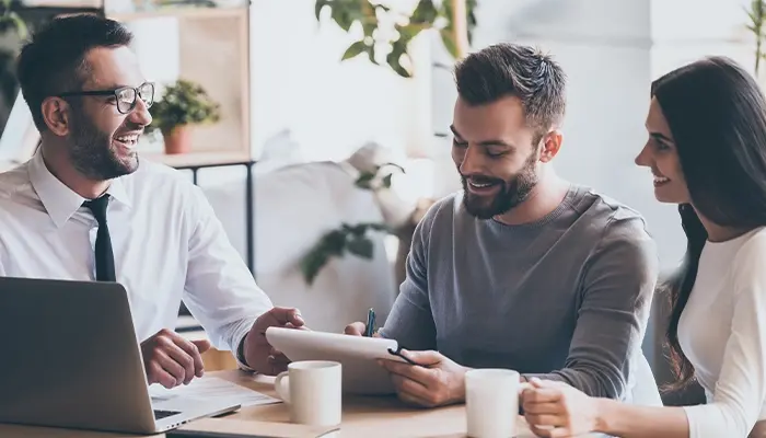 Three coworkers sit at a table in a bright office, smiling and discussing content on a tablet, with a laptop and coffee cups in front of them.